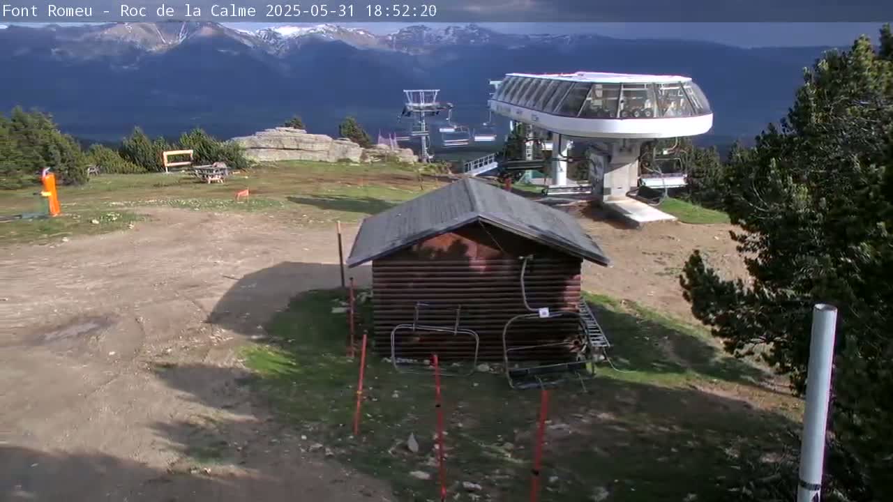 A small wooden cabin sits near a ski lift station on a partly sunny day with a mountain range in the background.