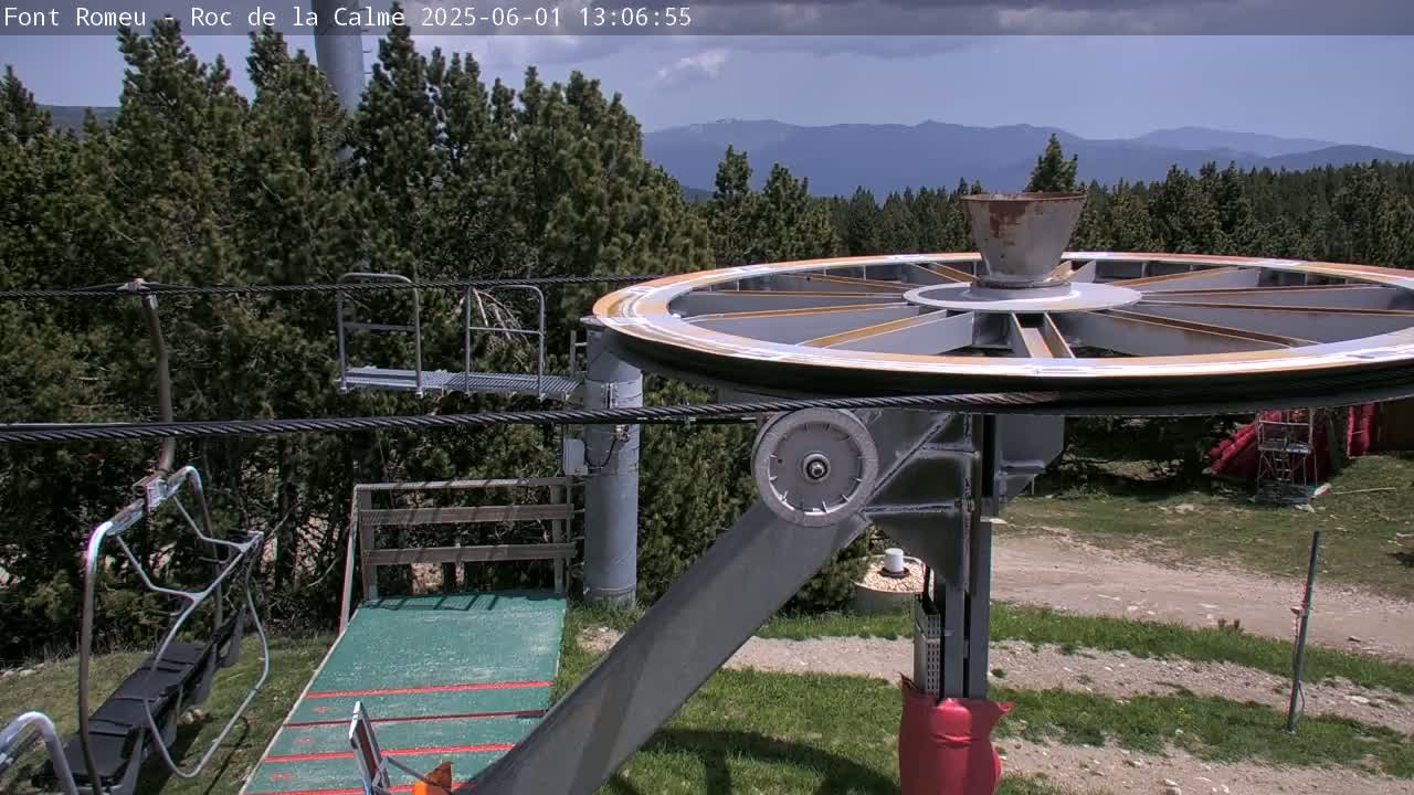 A large pulley system of a chairlift is seen in a mostly sunny area, with a mountainous backdrop and a forest of evergreen trees in the foreground.