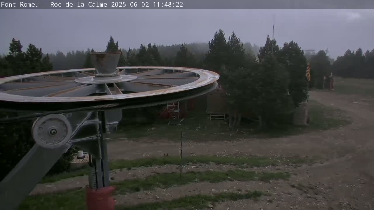 A large pulley wheel of a ski lift is in the foreground, with a view of a wooded area under an overcast sky.