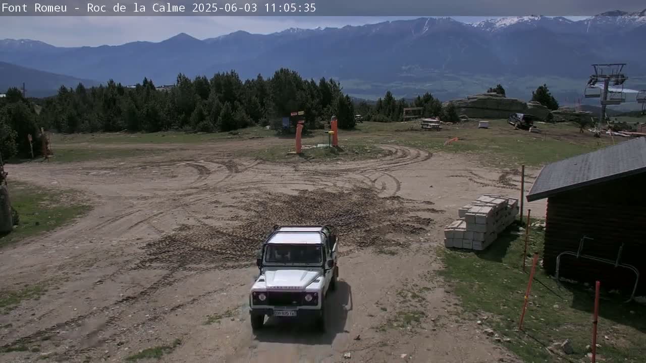 A white Land Rover drives on a dirt road in a mountainous area under a mostly clear sky, with a chairlift and a small building visible in the background.