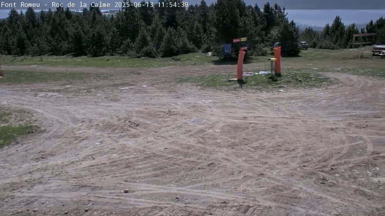 A mostly dirt area with tire tracks, sparse grass, and a few tall orange objects in the background, under a partly sunny sky.