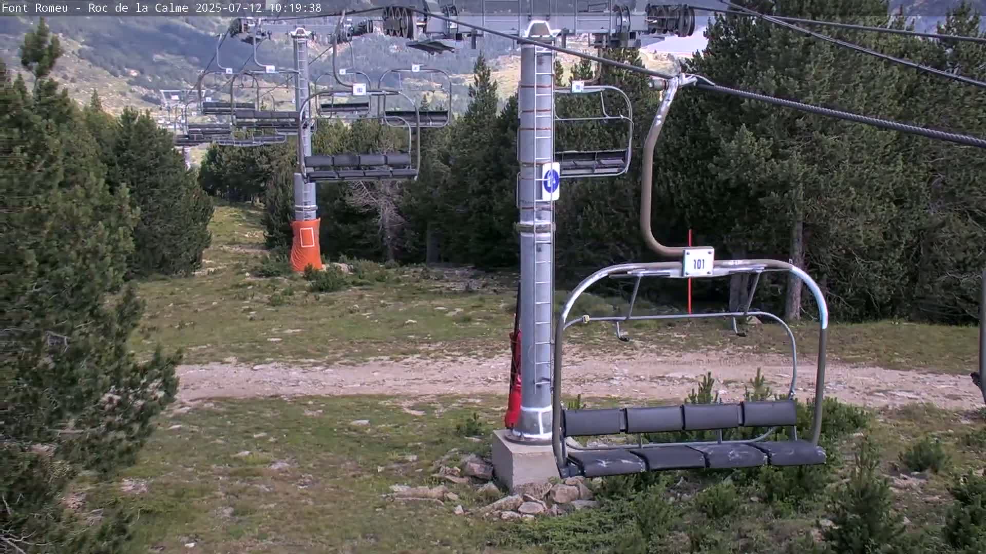 An empty chairlift hangs above a grassy, rocky area dotted with pine trees under a mostly clear sky.