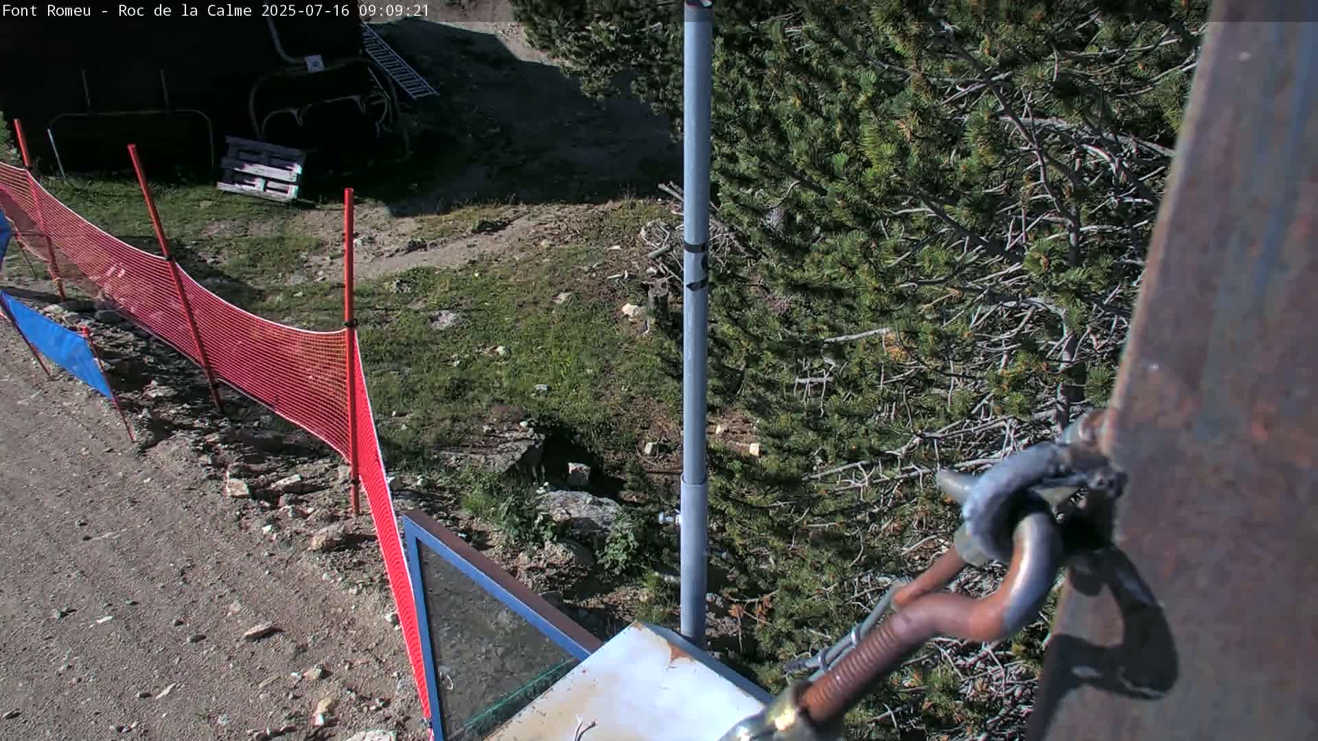 A view from a high angle shows a section of red and blue safety netting, a gray metal pole, a rusty metal hook and chain, and a green coniferous tree under sunny conditions.