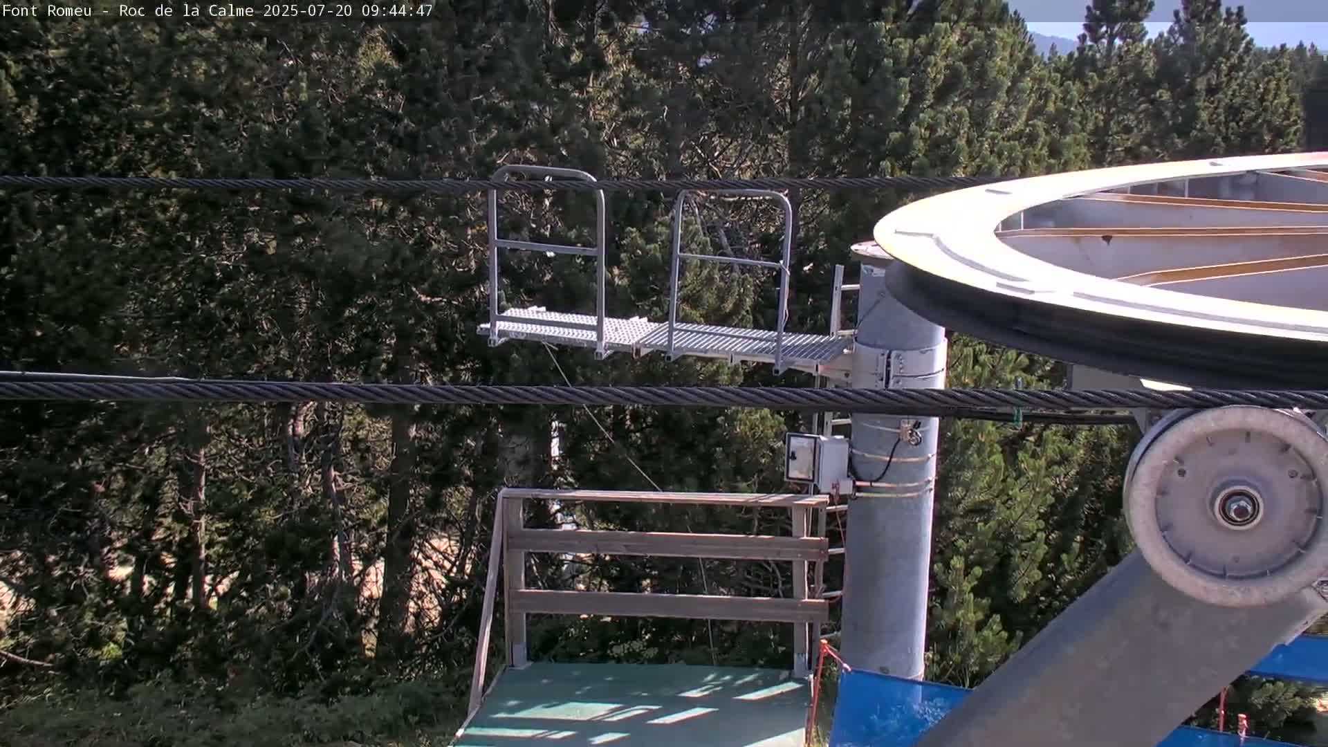 A chairlift mechanism is shown against a backdrop of a sunny, forested hillside.