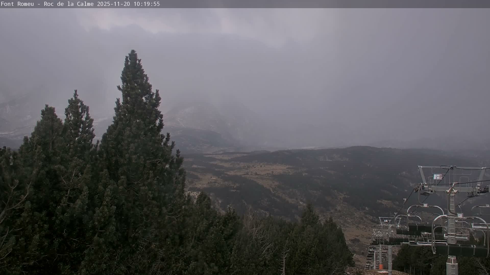 A mountain landscape is seen under heavily overcast and cloudy skies, with dense evergreen trees and a ski lift visible in the foreground overlooking distant valleys and peaks.