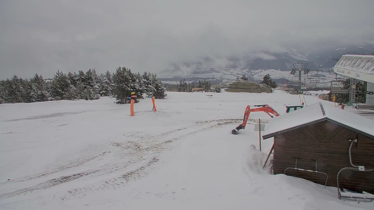 A mountain landscape is seen under heavily overcast and cloudy skies, with dense evergreen trees and a ski lift visible in the foreground overlooking distant valleys and peaks.