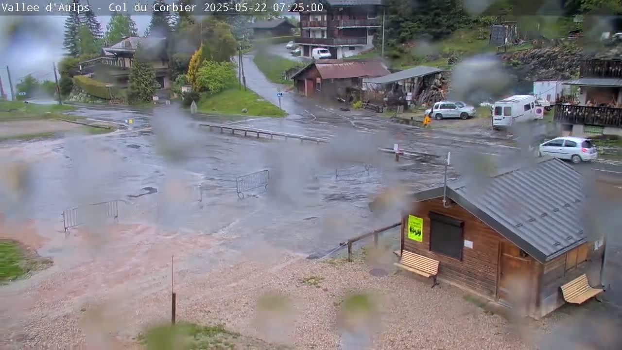 Floodwaters cover a roadway and parking area near a small wooden building and several vehicles, during a rainy day.