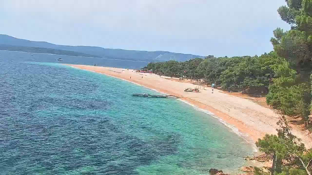 A long, sandy beach with shallow, turquoise water curves around a coastline backed by dark green trees under a clear sky.