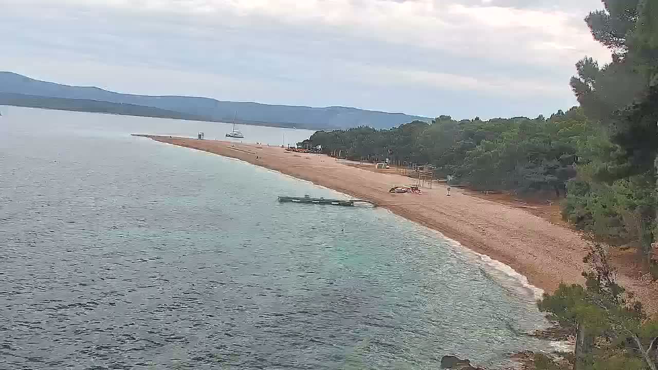 A long, sandy beach curves gently into calm, shallow blue water under an overcast sky, backed by dark green trees and distant mountains.