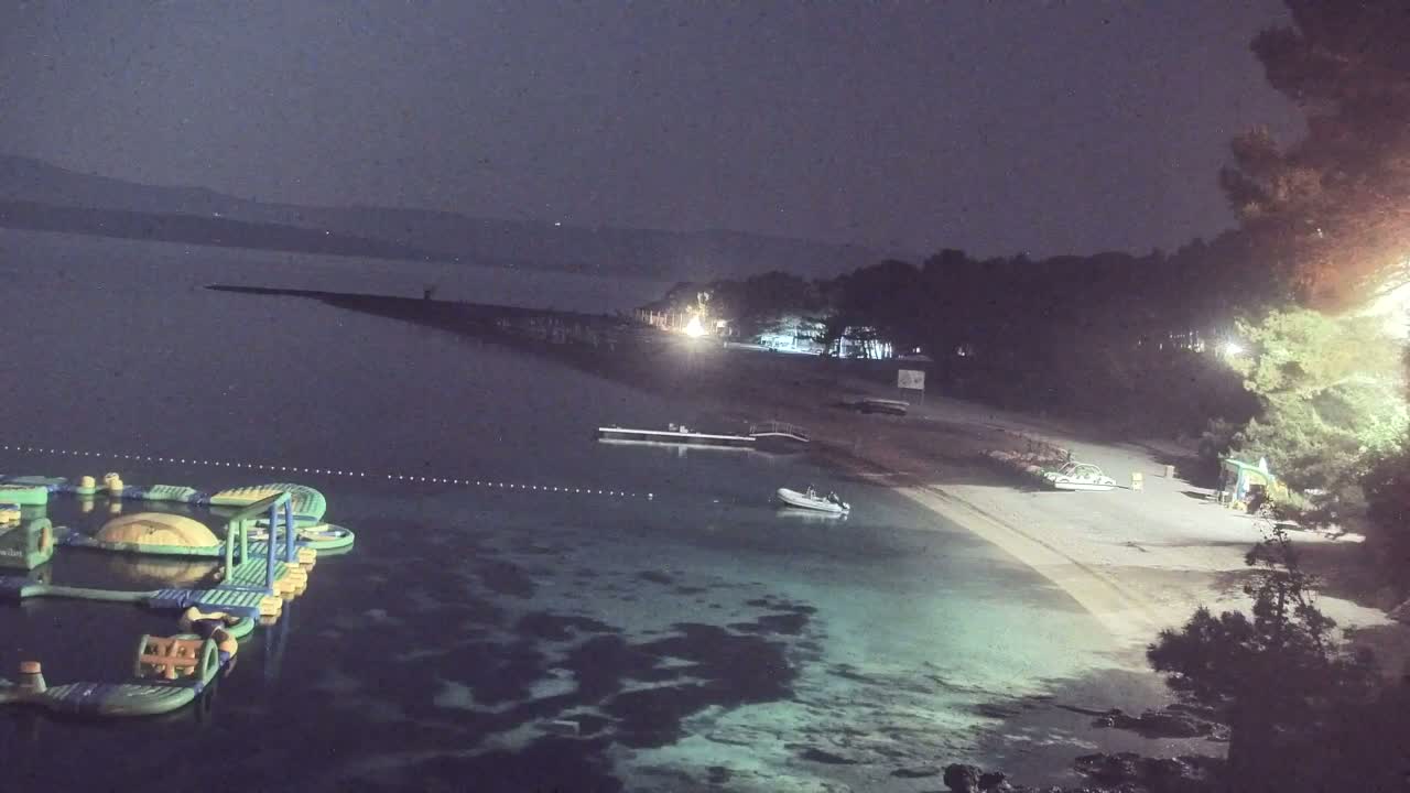 A nighttime view of a beach with calm, shallow water, an inflatable water park, a small boat, and trees illuminated by artificial lights.