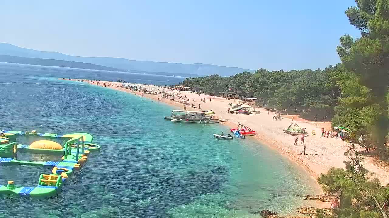 A long, sandy beach with clear, turquoise water, numerous people sunbathing and swimming, several boats anchored near the shore, and inflatable water games in the shallows, all under a sunny sky.