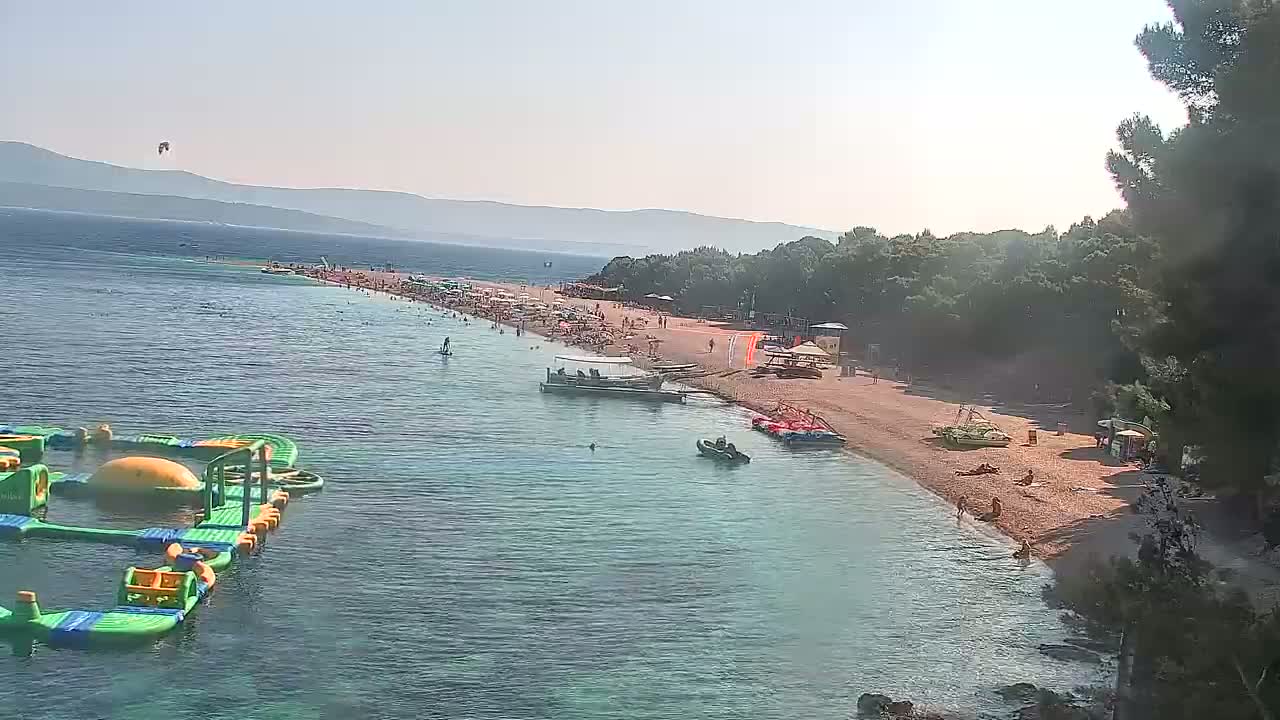 A long, sandy beach filled with people is bordered by calm, clear water and a line of green trees under a sunny sky, with a kitesurfer visible in the distance.