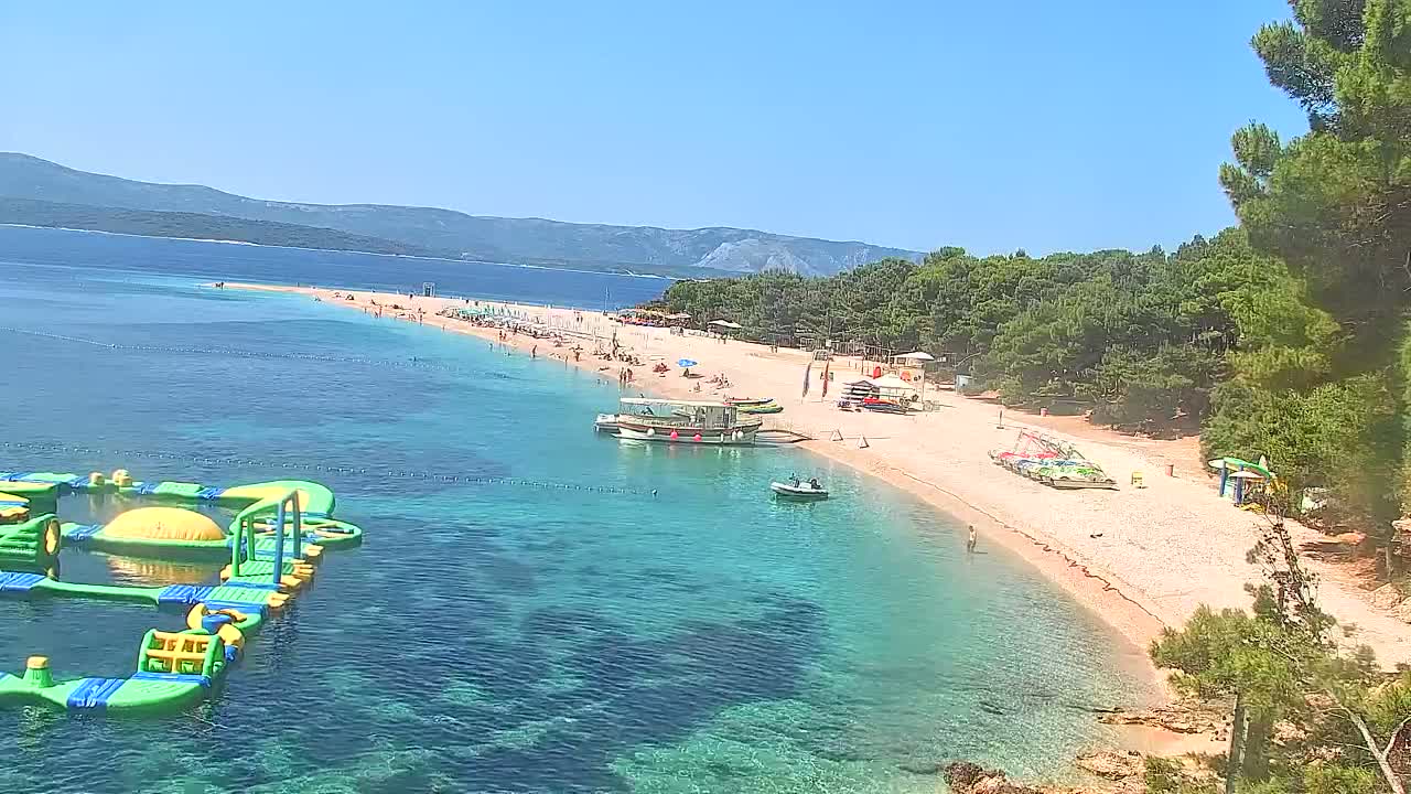 A long sandy beach with clear turquoise water, numerous beachgoers, several boats, and an inflatable water park is seen on a sunny day, with mountains visible in the background.