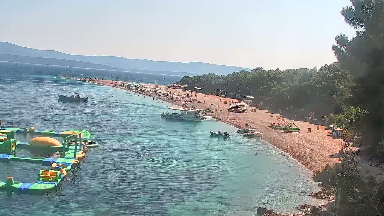 A sunny day at a beach shows a sandy shoreline with many people, several boats in the water, and inflatable water toys.