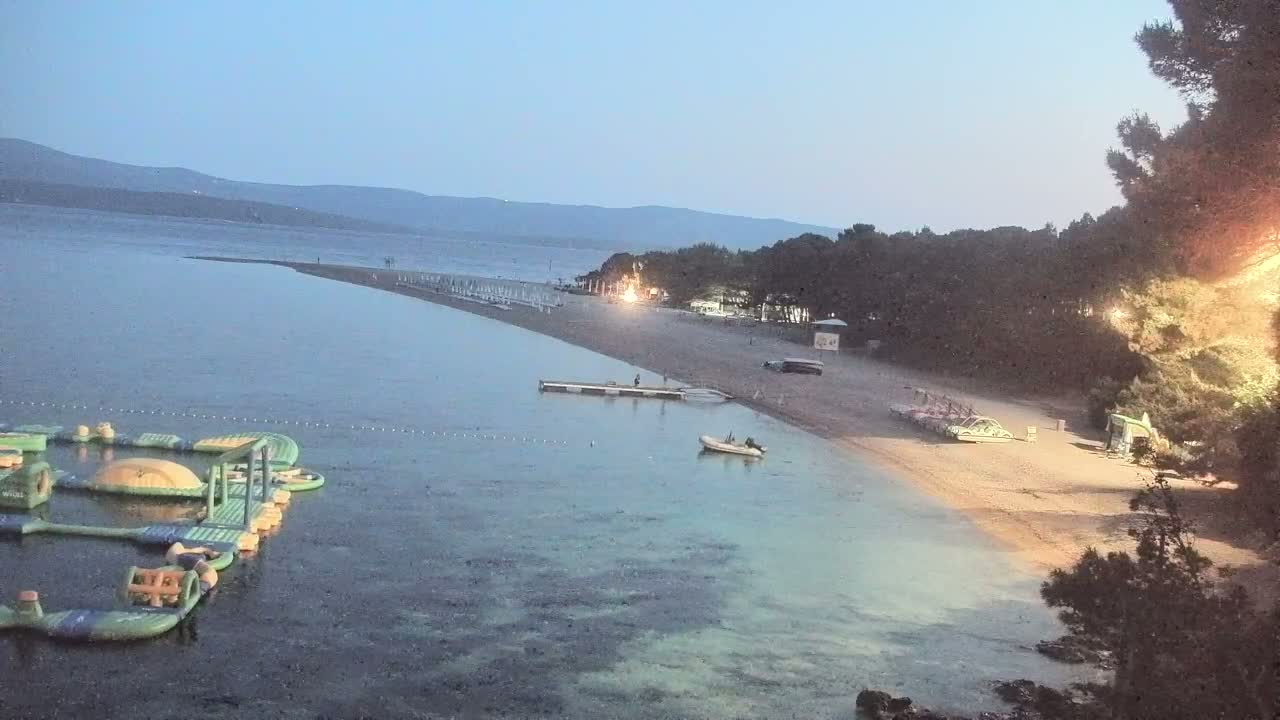 A sunny day at a beach shows a sandy shoreline with many people, several boats in the water, and inflatable water toys.