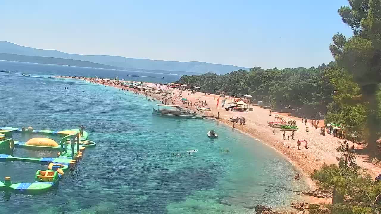 A long, sandy beach with numerous people on it and in the clear, shallow water, is backed by trees and mountains under a sunny sky.