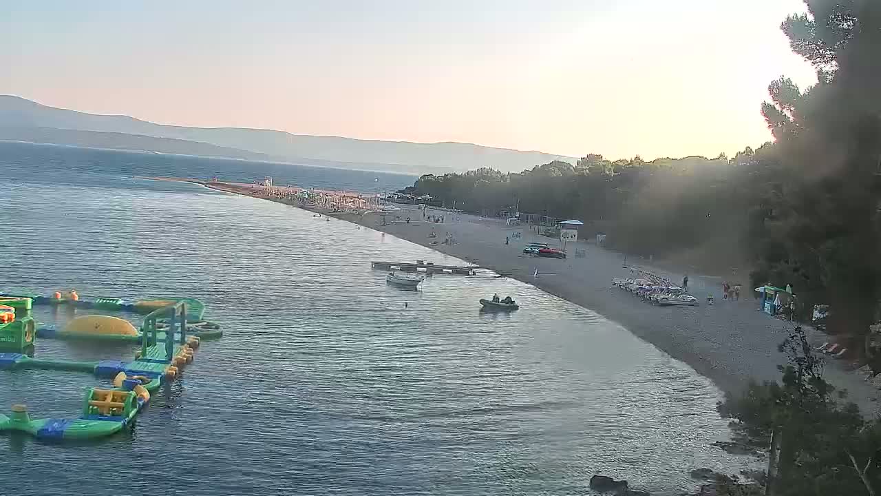 An aerial view of a beach scene at sunset, showing calm water, a sandy shore with people, several small boats, and an inflatable water park.