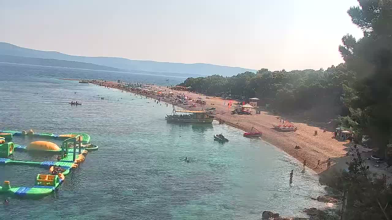 A sunny day at a beach shows a sandy shoreline with many people swimming and sunbathing, two boats docked near the shore, an inflatable water park, and a background of green hills and clear blue water.