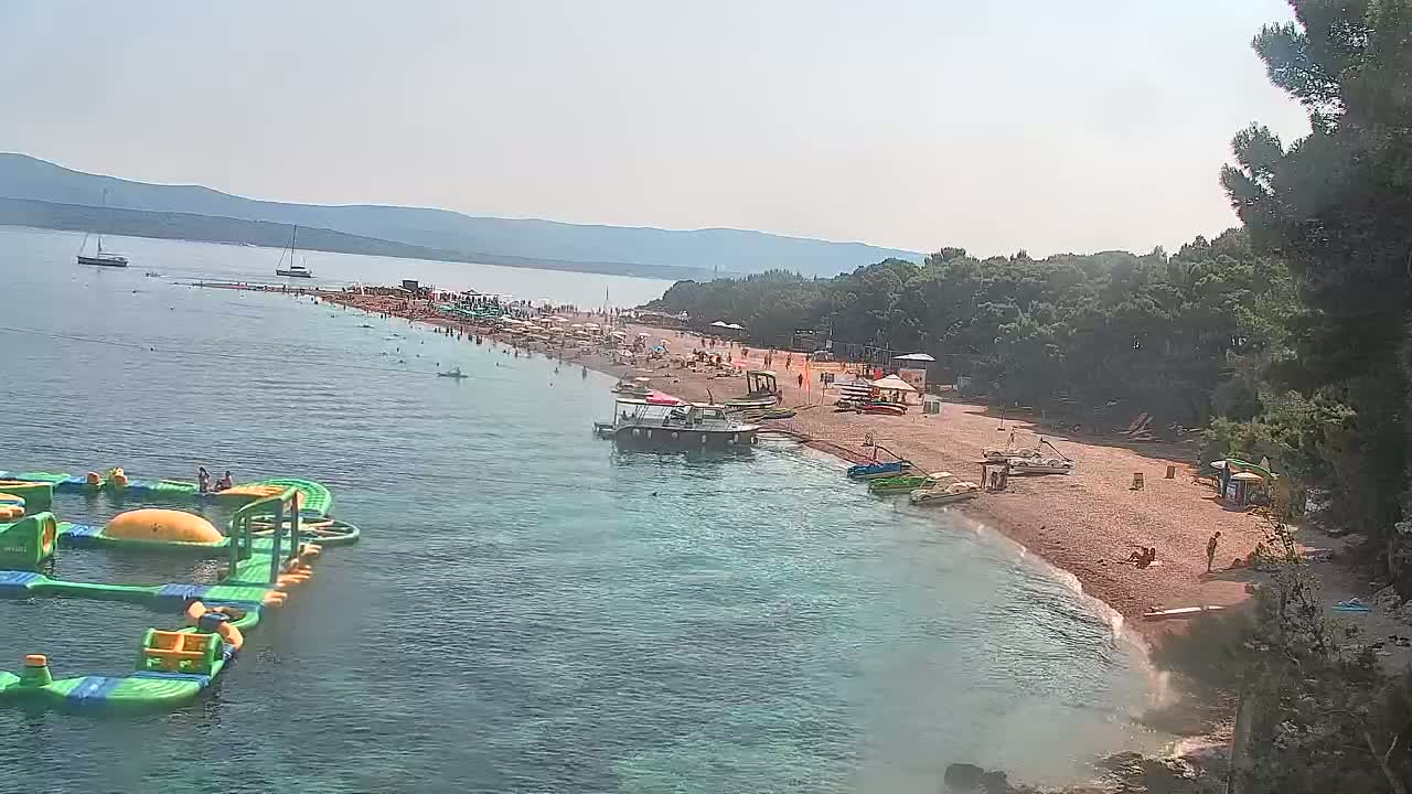 A crowded beach with clear, shallow water, an inflatable water park, several boats, and a background of green hills under a sunny sky.