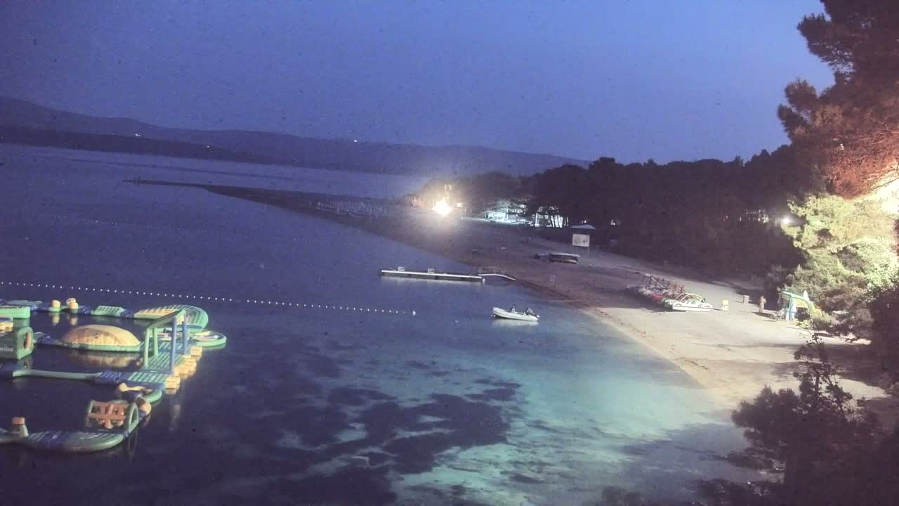 An inflatable water park is in calm, shallow, twilight-lit water near a sandy beach and shoreline trees under a dark blue night sky.