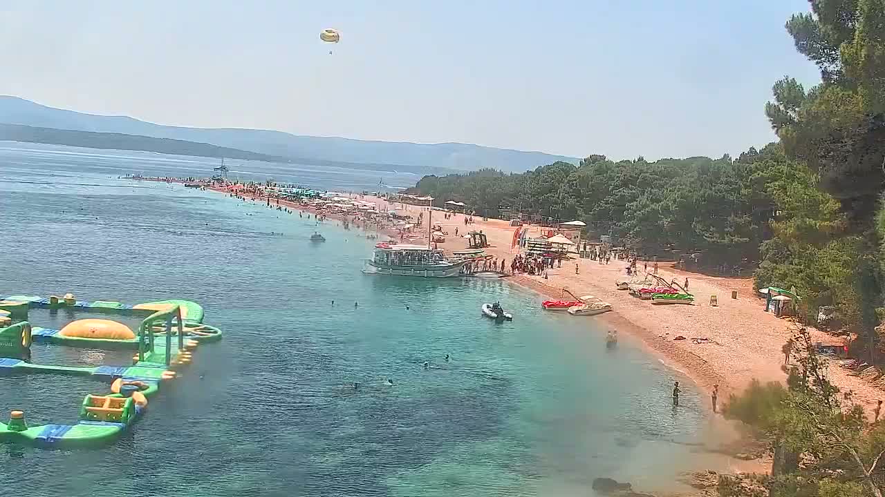 A sunny day at a crowded beach with clear turquoise water, a boat docked near the shore, and an inflatable water park in the sea.