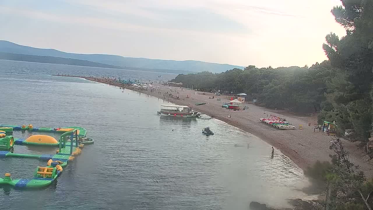 A long, sandy beach with many people on it, a boat offshore, and an inflatable water park in the calm sea under a partly cloudy sky.