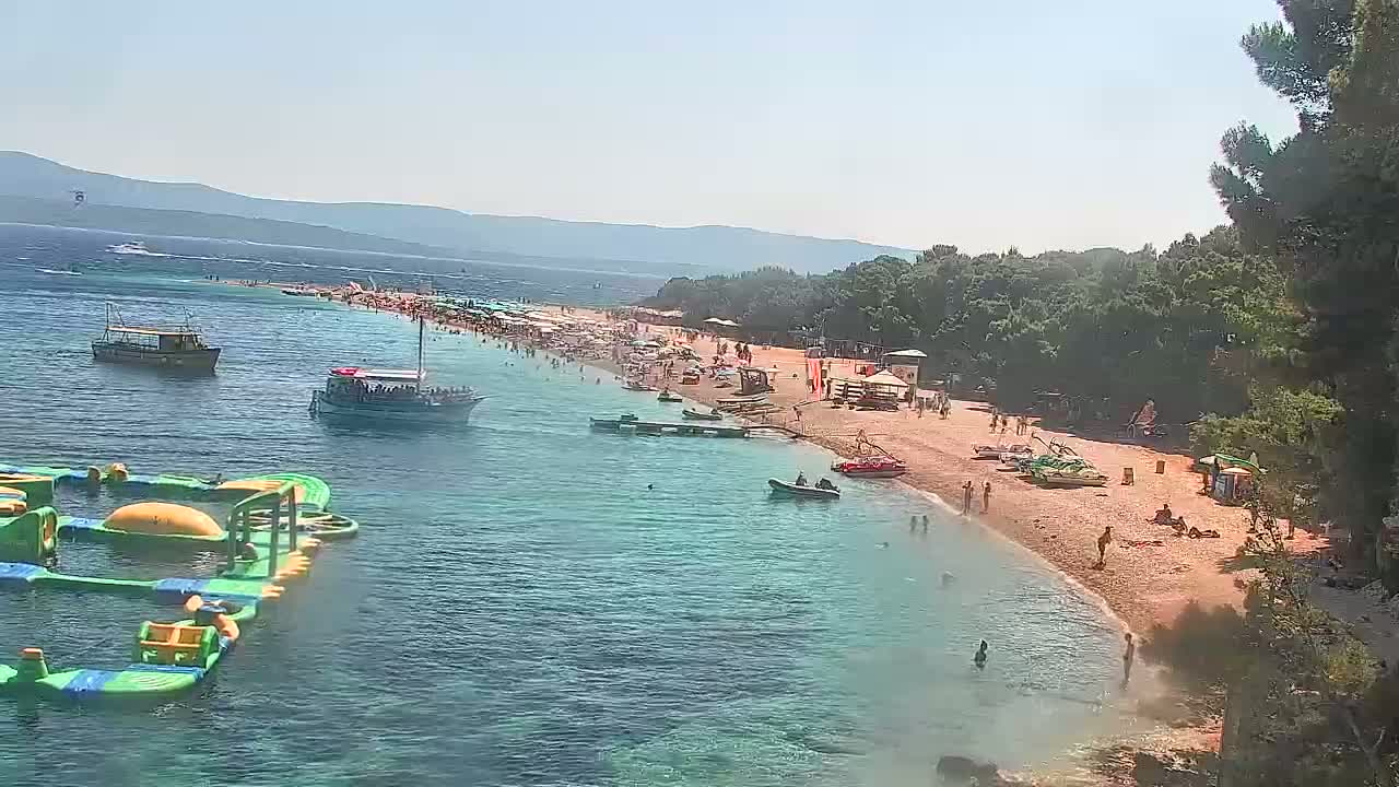 A crowded beach with turquoise water, several boats, and an inflatable water park is seen on a sunny day, with mountains in the background.