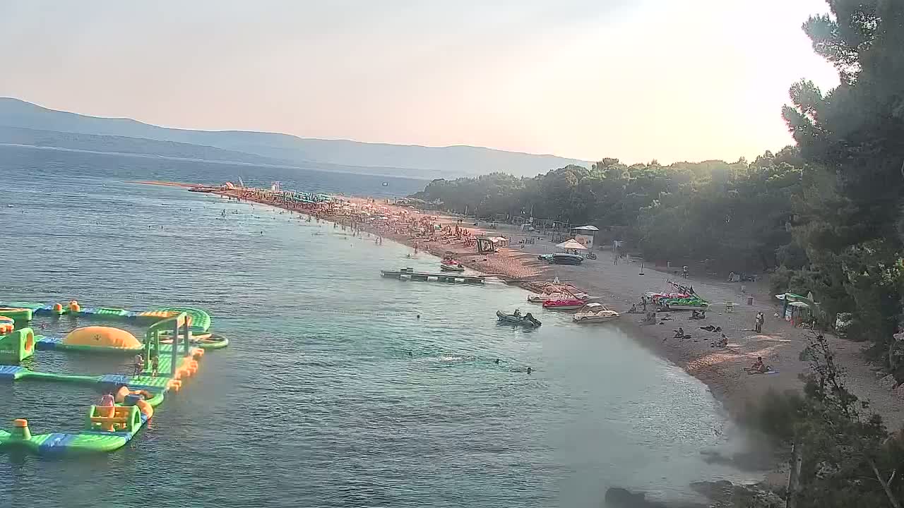 A crowded beach with turquoise water, inflatable water park, and boats is situated beneath a partly sunny sky in front of a line of green trees and distant mountains.