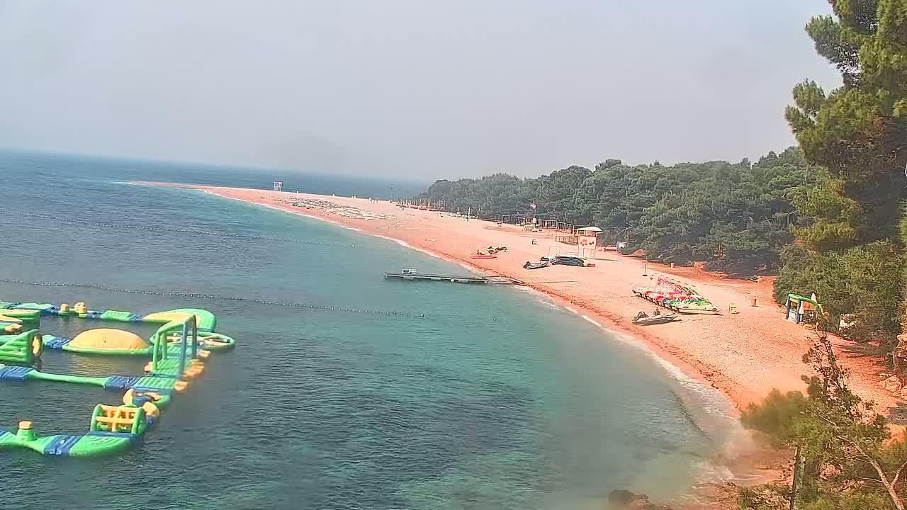 A hazy day shows a sandy beach with a line of trees behind it,  a small dock, several watercraft, and an inflatable water park in the calm sea.