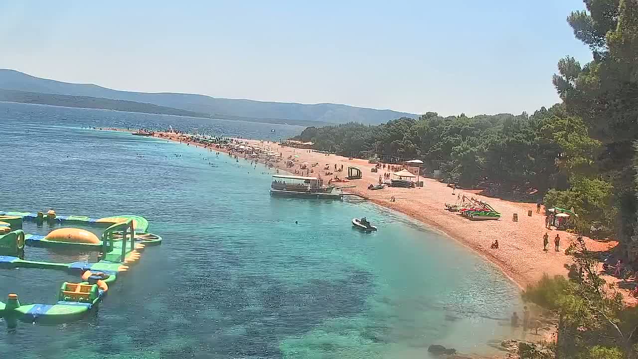 A long, sandy beach with many people sunbathing and swimming in clear, turquoise water, under a sunny sky, is bordered by green trees and mountains in the distance.