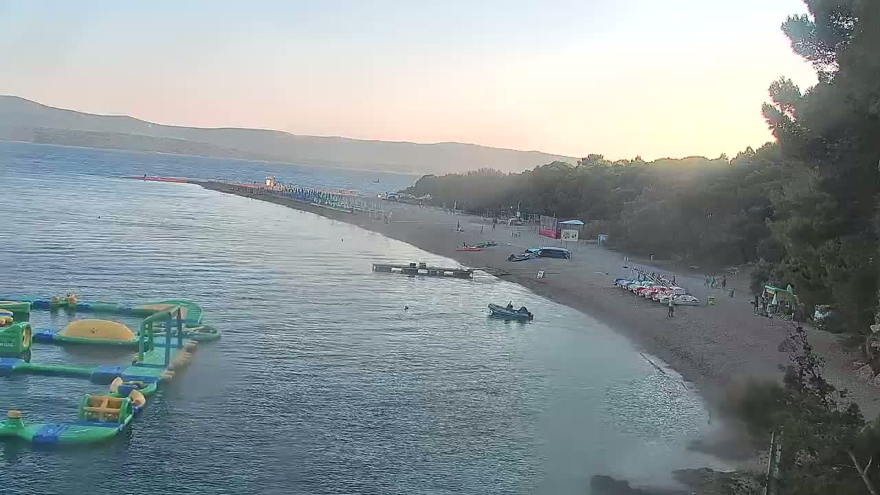 An aerial view shows a calm, sunlit beach with people, small boats, and an inflatable water park, backed by trees and mountains under a clear evening sky.