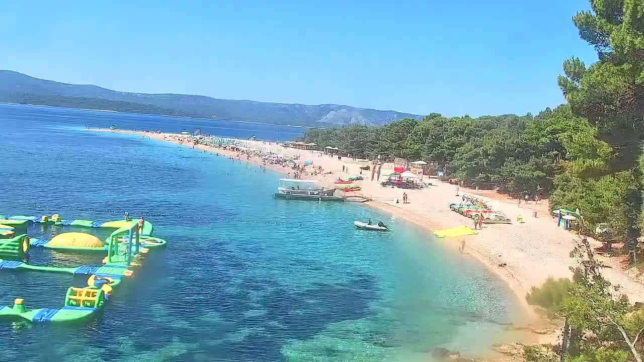A sunny day at a sandy beach with clear turquoise water, featuring numerous people, several small boats, and an inflatable water park.