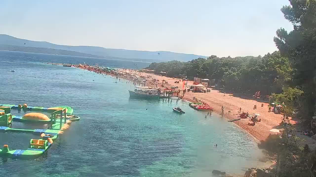 A sunny day at a crowded beach with clear turquoise water, a boat docked near the shore, and inflatable water park equipment in the water.