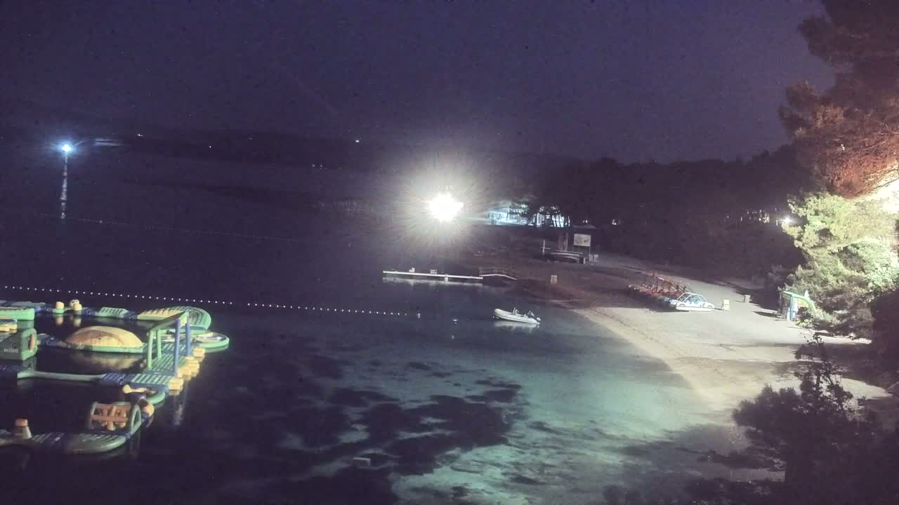 A nighttime view of a calm body of water with inflatable water park equipment, a small boat, and a sandy beach bordered by trees under a dark sky.