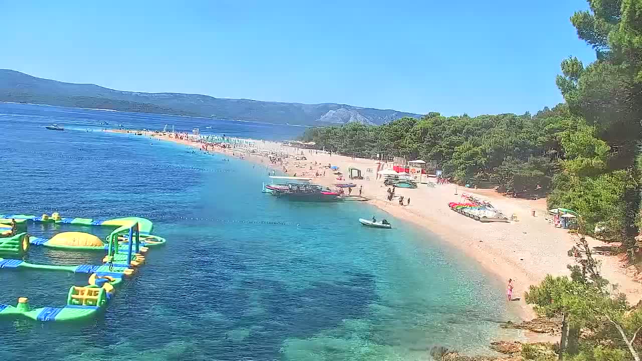 A sunny day at a beach shows a long sandy shore with many people, water toys in the ocean, boats docked near the beach, and green trees behind the beach.