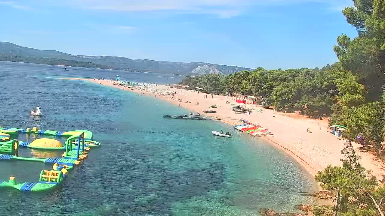 A sandy beach with clear, shallow turquoise water, populated with people and watercraft, is backed by lush green vegetation and mountains under sunny skies.