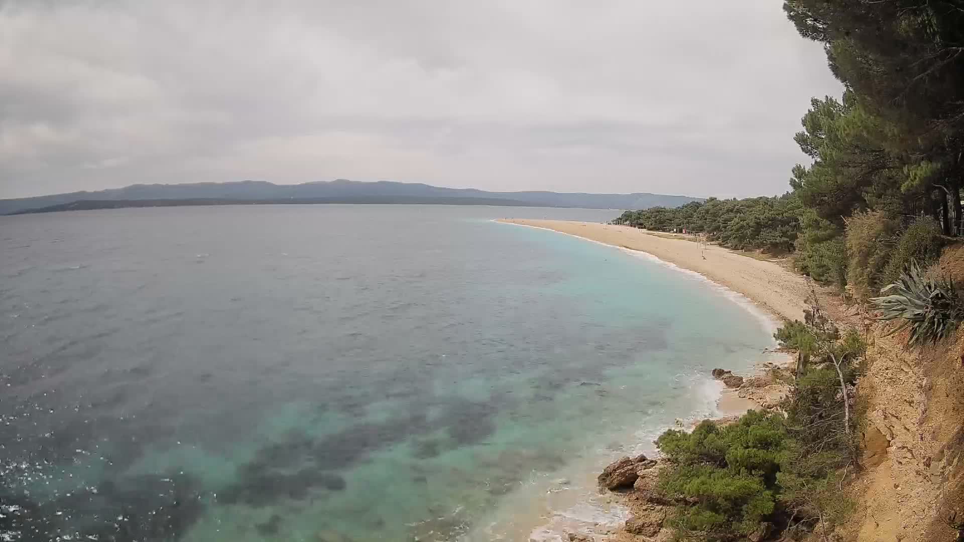 A sandy beach with clear, shallow turquoise water, populated with people and watercraft, is backed by lush green vegetation and mountains under sunny skies.