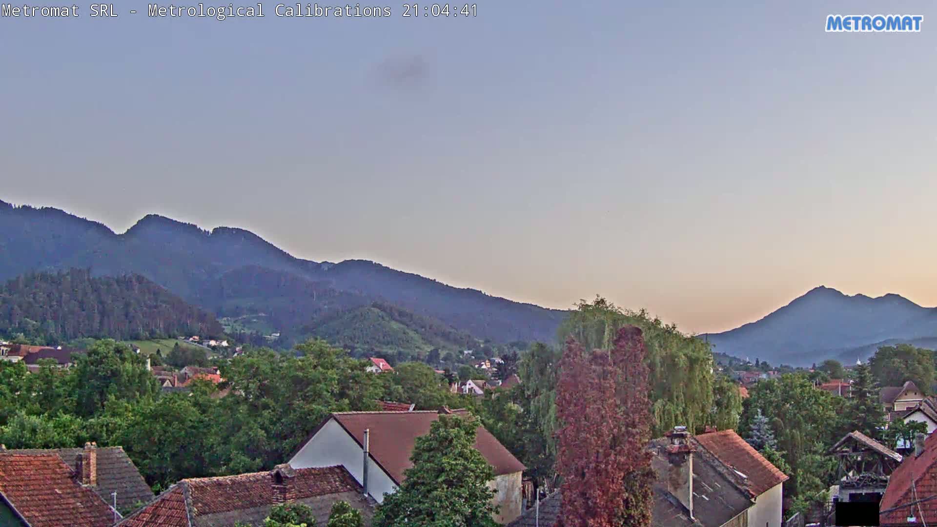 A twilight view shows a village nestled among green trees and hills, with mountains in the background under a clear, light-purple sky.