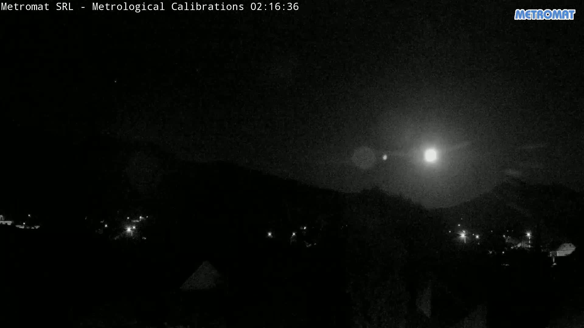 A nighttime, low-light view of a town or village nestled at the base of a hill or mountain, illuminated by a bright moon and a few scattered lights.