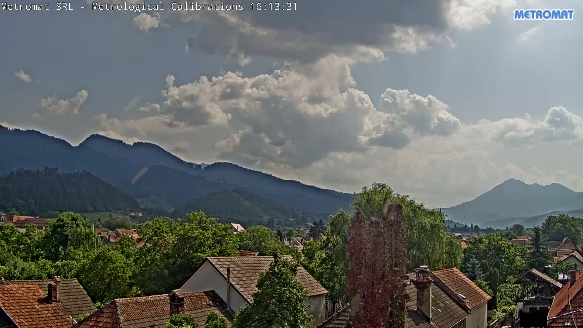 A partly cloudy sky overlooks a village nestled among green, tree-covered hills and mountains.