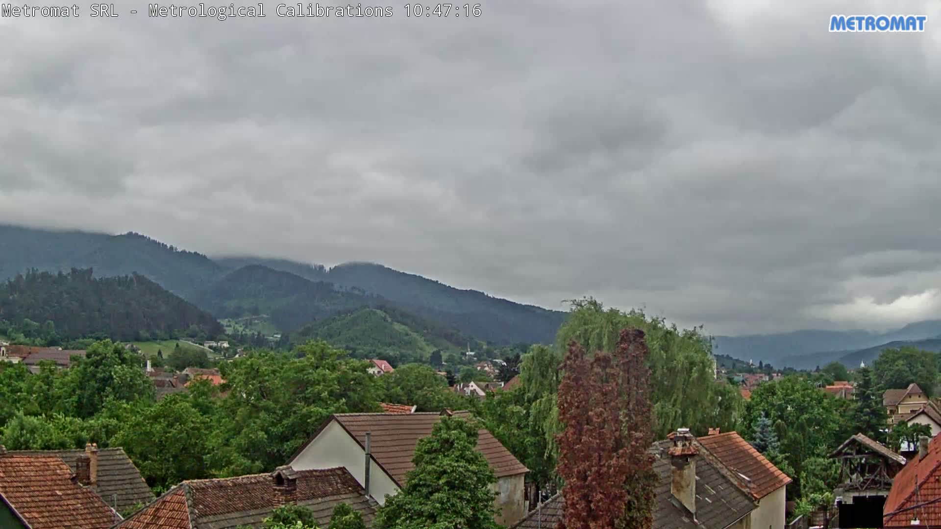 Overcast skies loom over a village nestled among green, tree-covered hills and mountains.