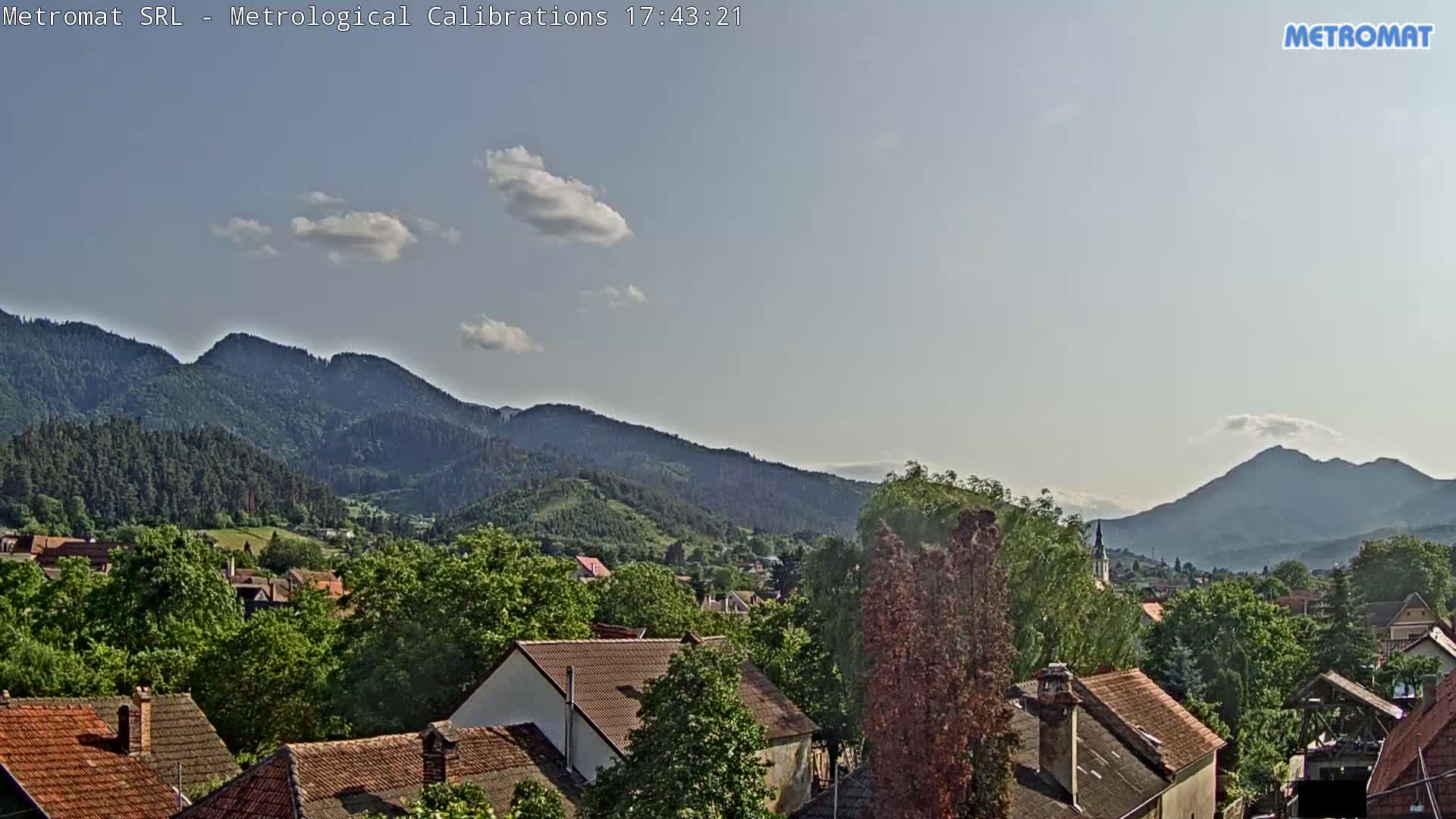 A mostly sunny view shows a village nestled among green-covered hills and mountains under a mostly clear sky.