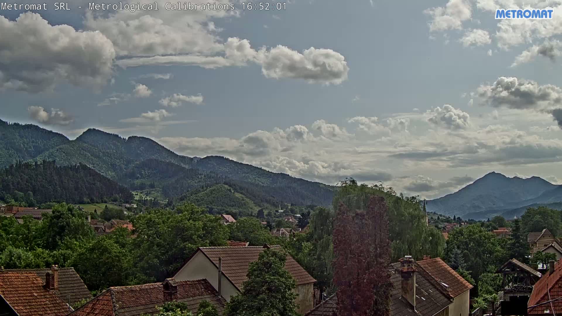 A partly cloudy sky overlooks a village nestled among green, tree-covered mountains.