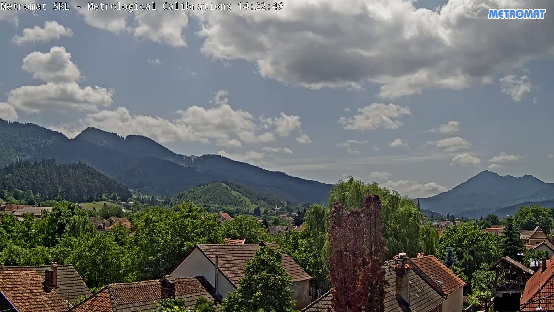 A partly cloudy day reveals a village nestled among lush green trees and rolling hills, with mountains visible in the background.
