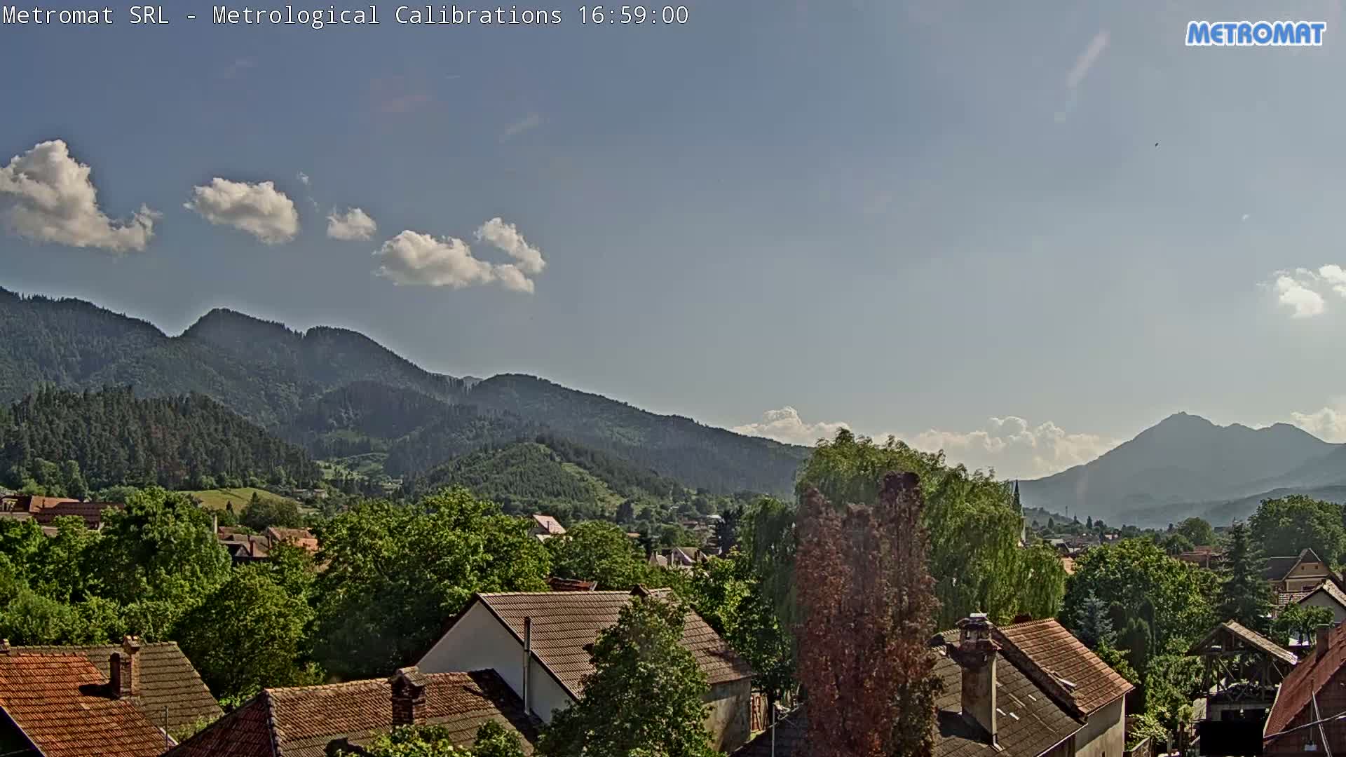 A partly sunny day reveals a village nestled among green, tree-covered hills and mountains.