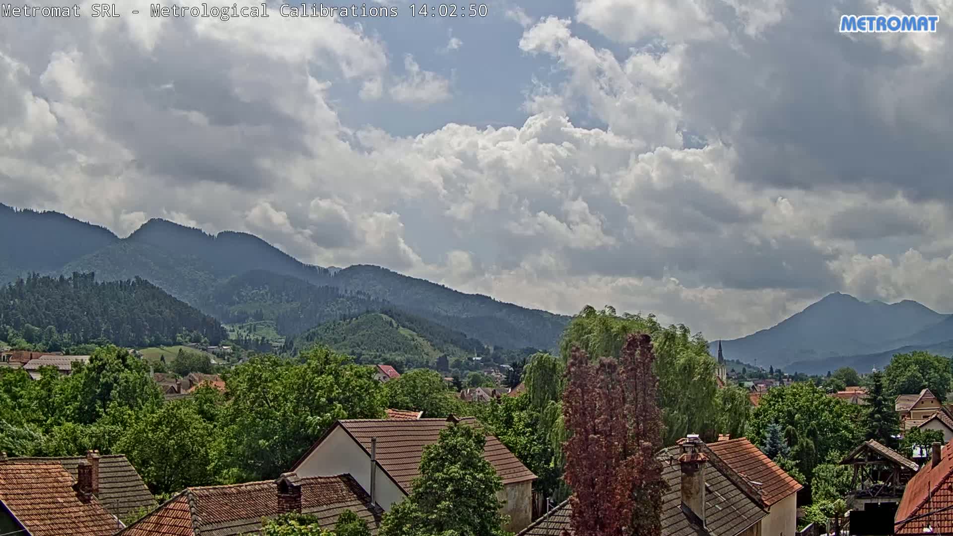 A partly cloudy sky overlooks a village nestled among green, forested mountains.