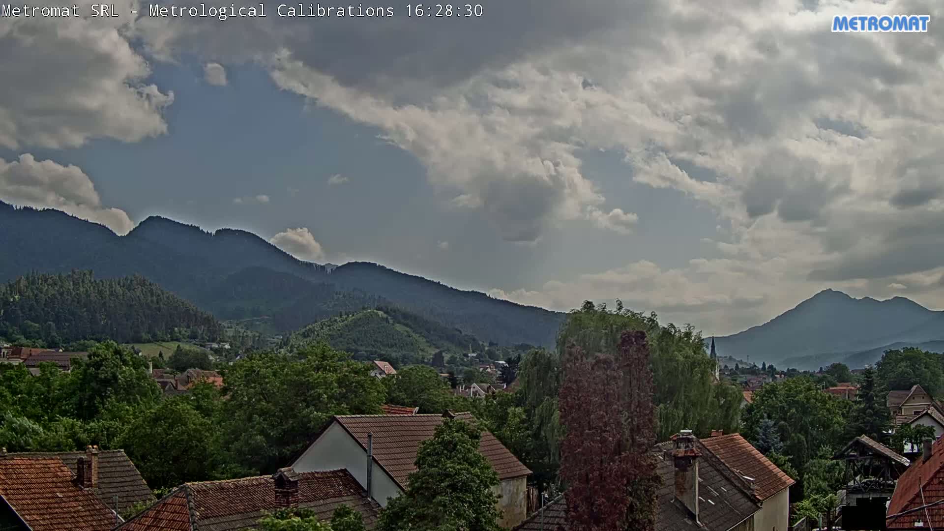 A partly cloudy sky overlooks a village nestled among green, tree-covered hills and mountains.