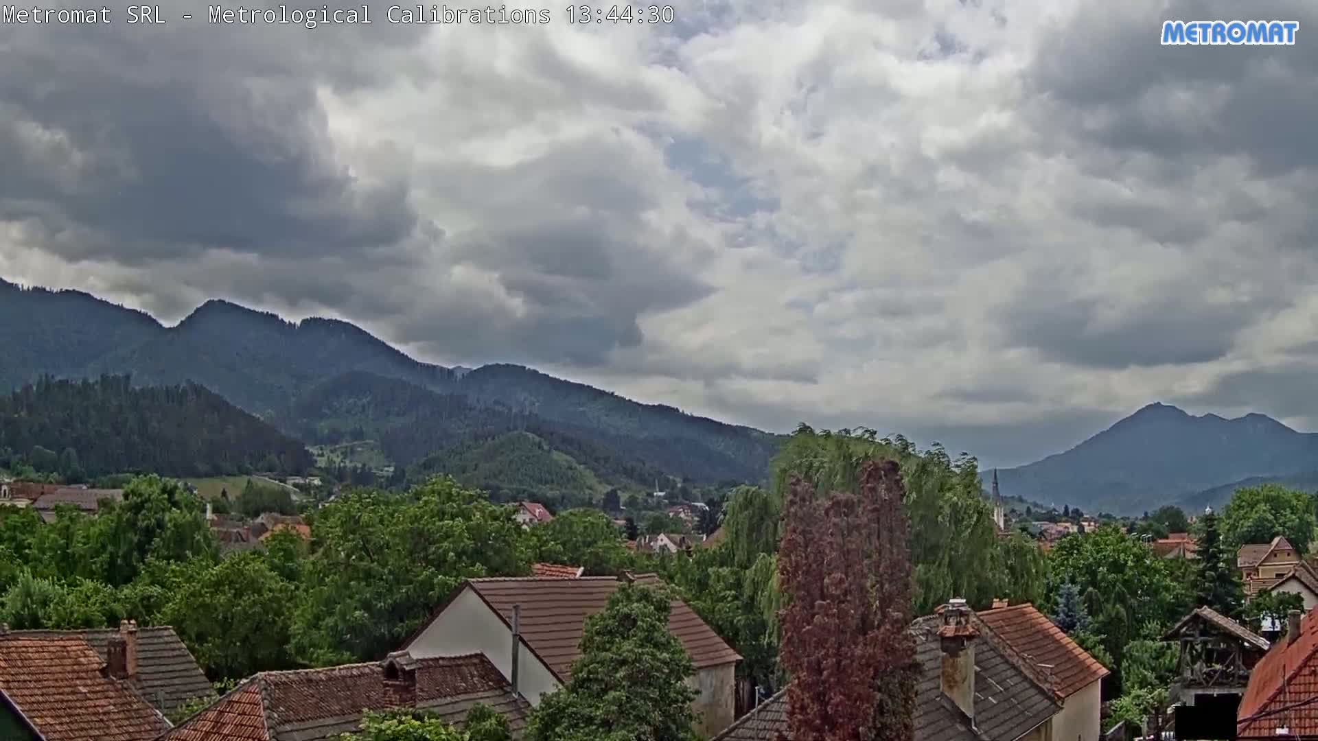 A view of a town nestled in a valley, surrounded by green hills and mountains under a mostly cloudy sky.