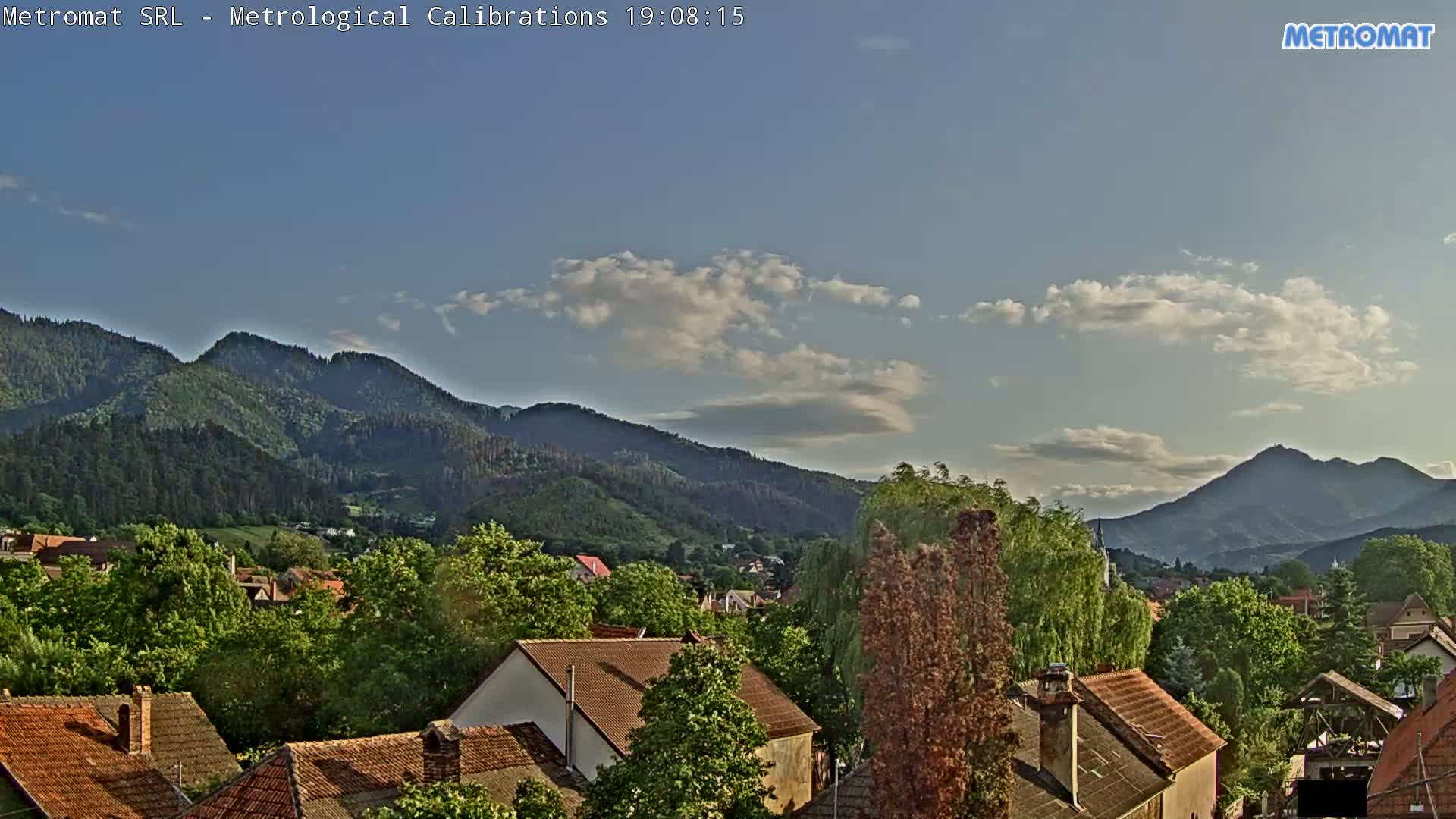 A partly cloudy sky overlooks a village nestled among green, tree-covered mountains.