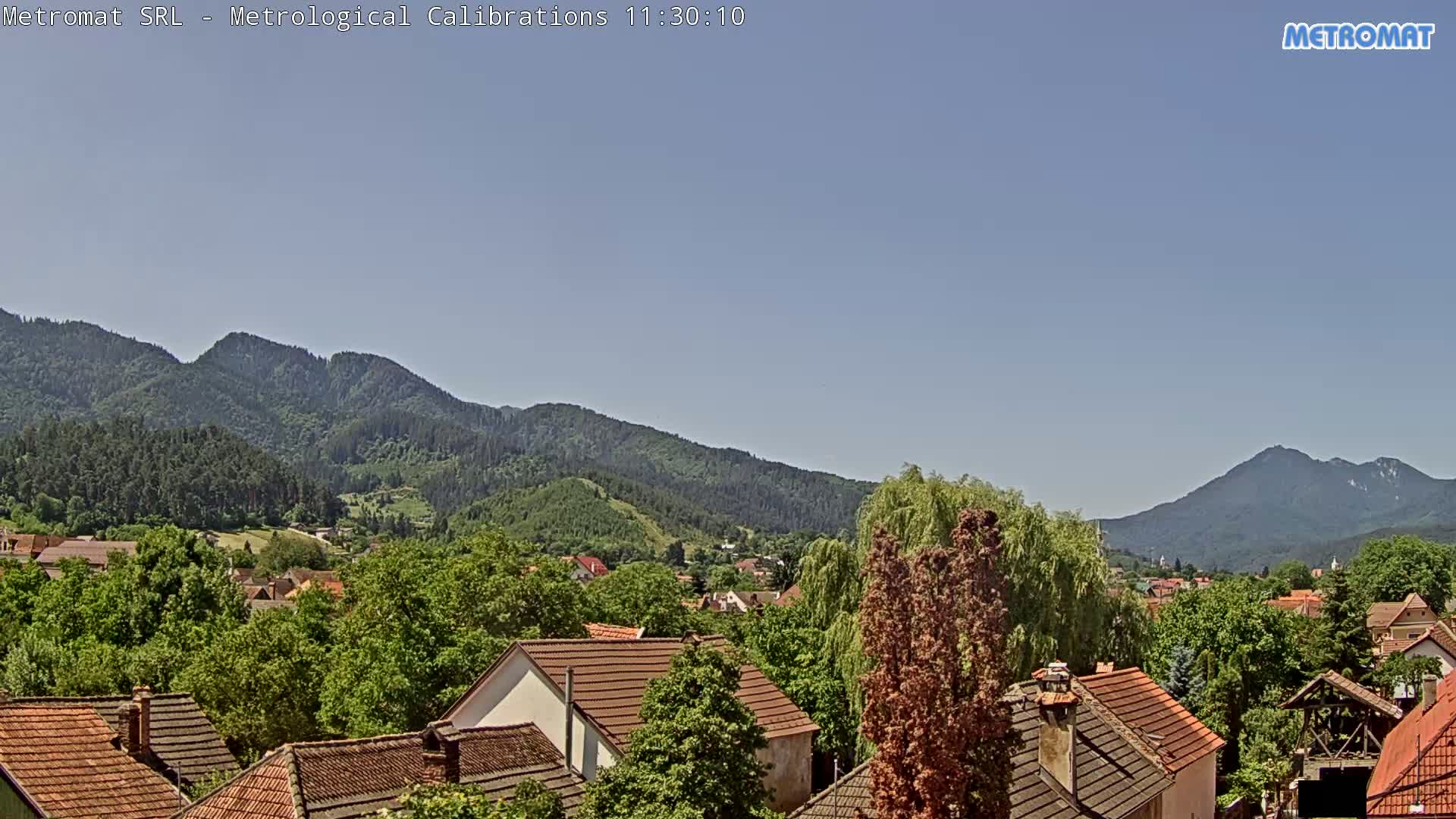 A clear, sunny day reveals a village nestled amongst lush green trees and mountains.